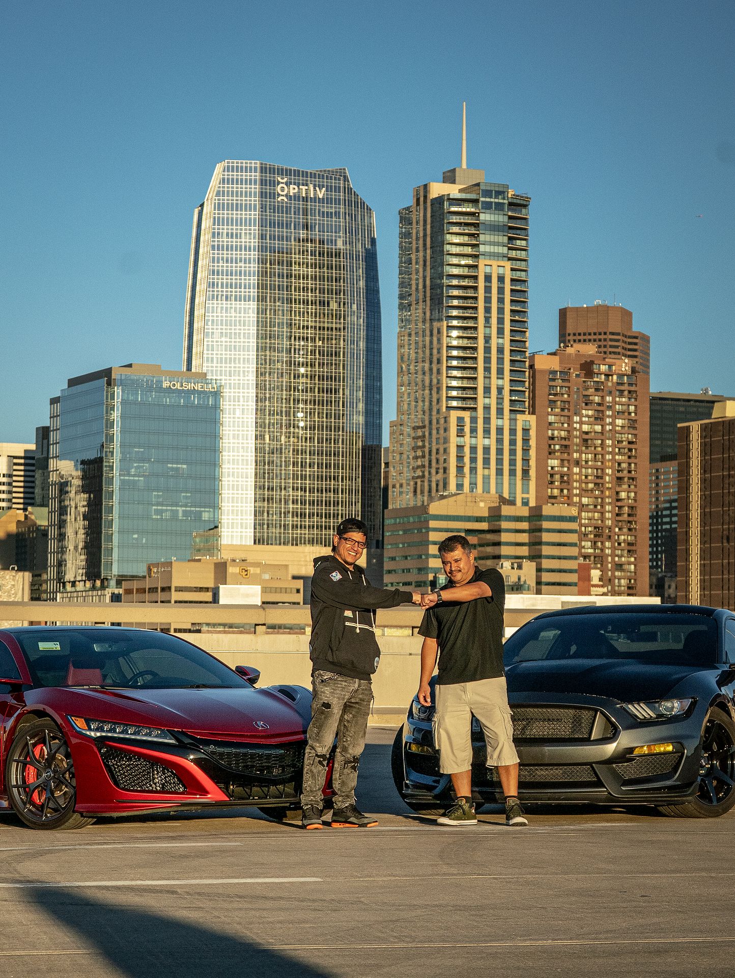 Two owners of a Grey 2018 Ford Mustang Shelby and a Red 2021 Acura NSX bumping fists while standing in front of their vehicles with a downtown Denver background